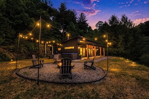 A cabin with a deck, surrounded by trees, and string lights. - Brand New Luxe Modern Cabin: Views, Hot tub, Creek (Ellijay)