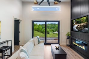 A modern living room with a sofa, a coffee table, a fireplace, and a large window offering a view of a green landscape. - Brand New Luxe Modern Cabin: Views, Hot tub, Creek (Ellijay)