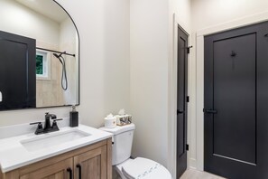 Une salle de bain avec un lavabo blanc, des toilettes, une porte noire et une fenêtre donnant sur un paysage verdoyant.