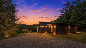 Une cabane moderne avec une allée en gravier, entourée d’arbres et sous un ciel dégagé.