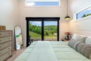 Une chambre avec un grand lit, une commode, un miroir, une table de chevet ornée d’un vase, et une vue sur un paysage verdoyant par le biais d’une porte coulissante.