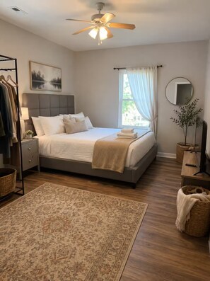 Ein Schlafzimmer mit Bett, Deckenventilator, Fenster mit Vorhängen, Nachttisch, Spiegel, Pflanze und Stehlampe.