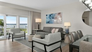 A modern hotel room with a balcony, a grey sofa set, two armchairs, a coffee table, and a dining table with chairs.
