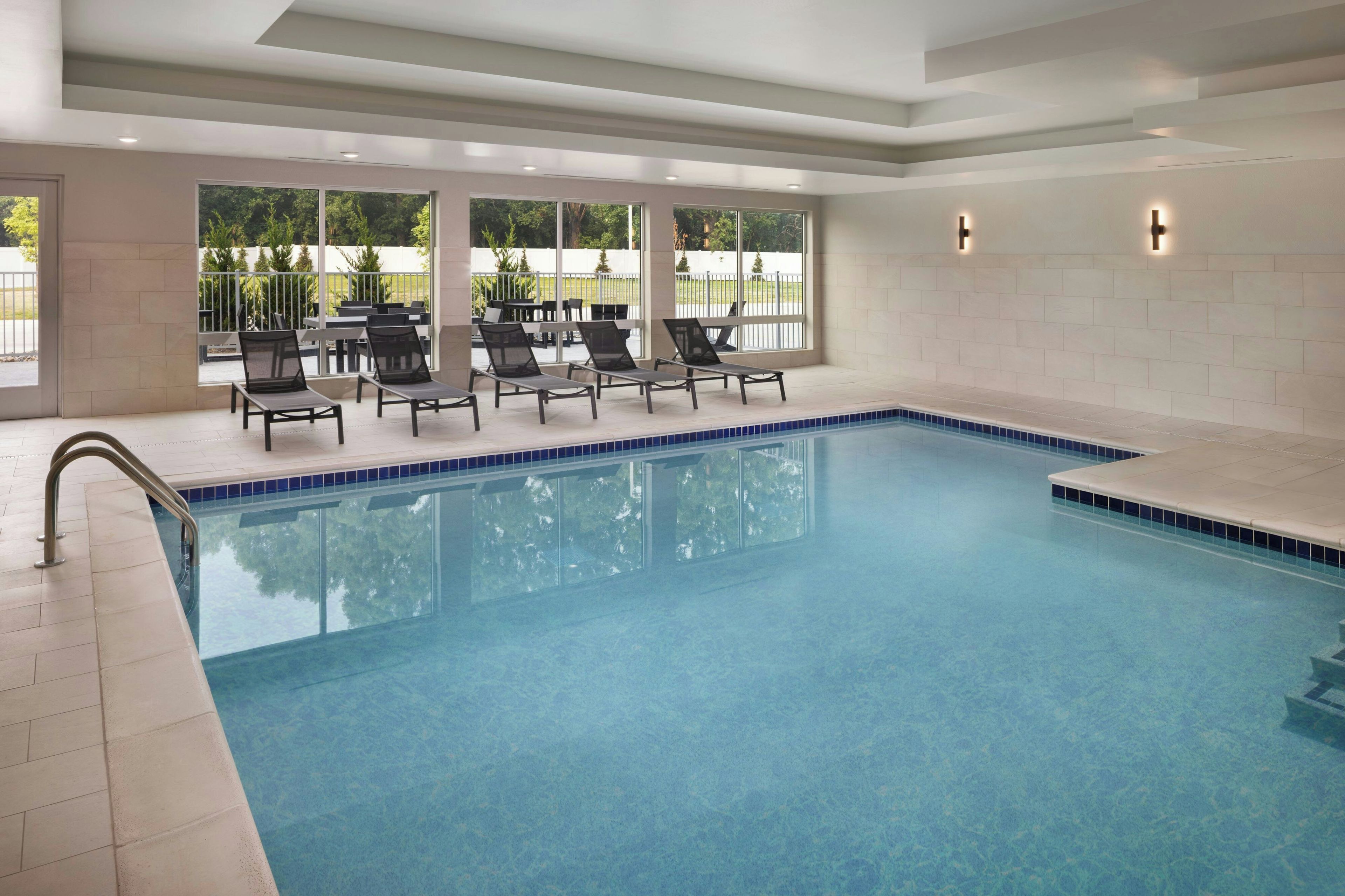 An indoor swimming pool with clear blue water, surrounded by lounge chairs and a view of greenery outside.
