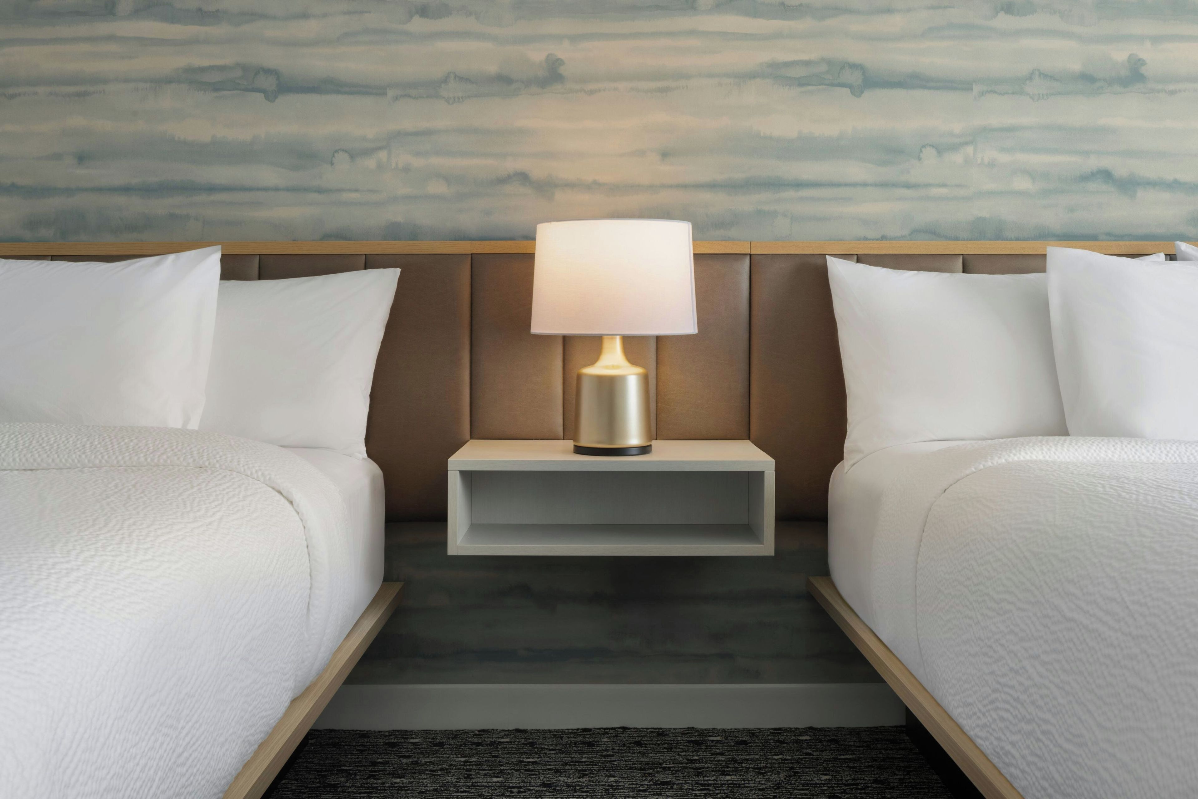 A hotel room with two beds, a nightstand with a lamp, and a wall with a painted landscape.