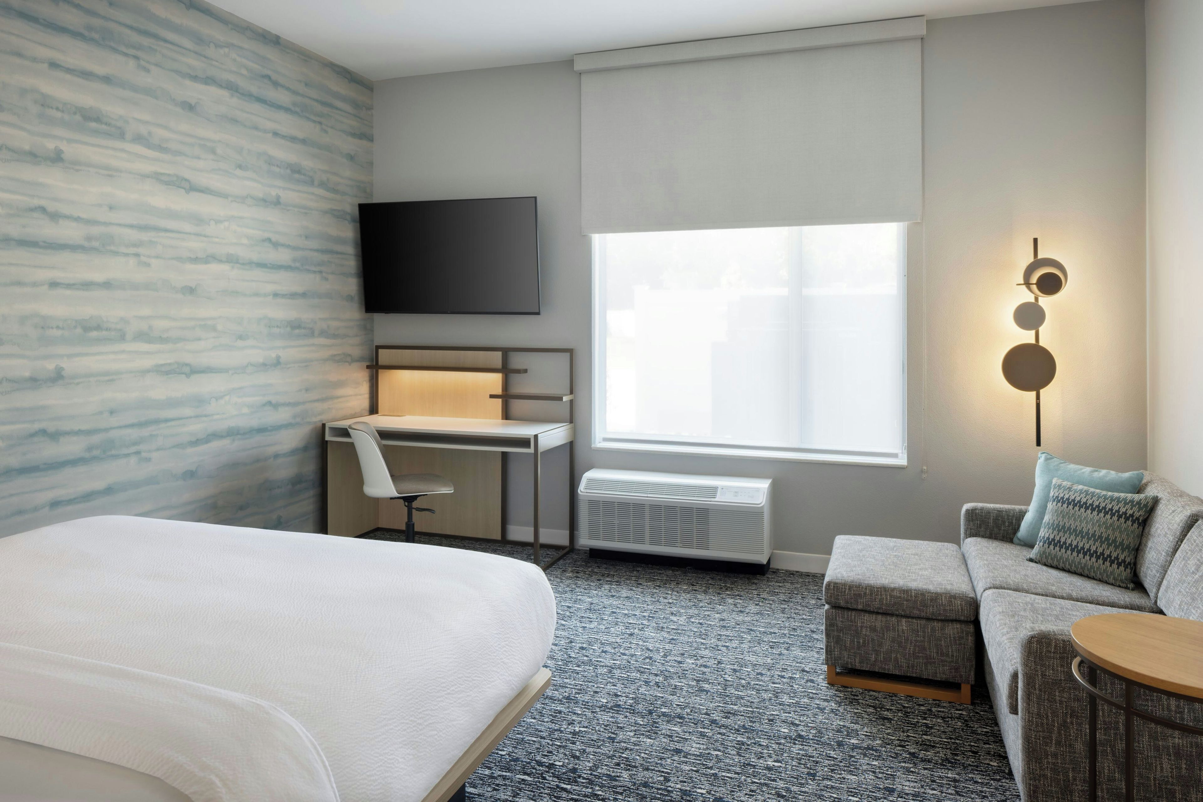Une chambre d’hôtel comprenant un lit, un bureau avec une chaise, une télévision, une fenêtre avec des stores, un radiateur et un canapé avec un coussin.
