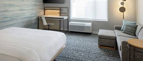 Une chambre d’hôtel comprenant un lit, un bureau avec une chaise, une télévision, une fenêtre avec des stores, un radiateur et un canapé avec un coussin.