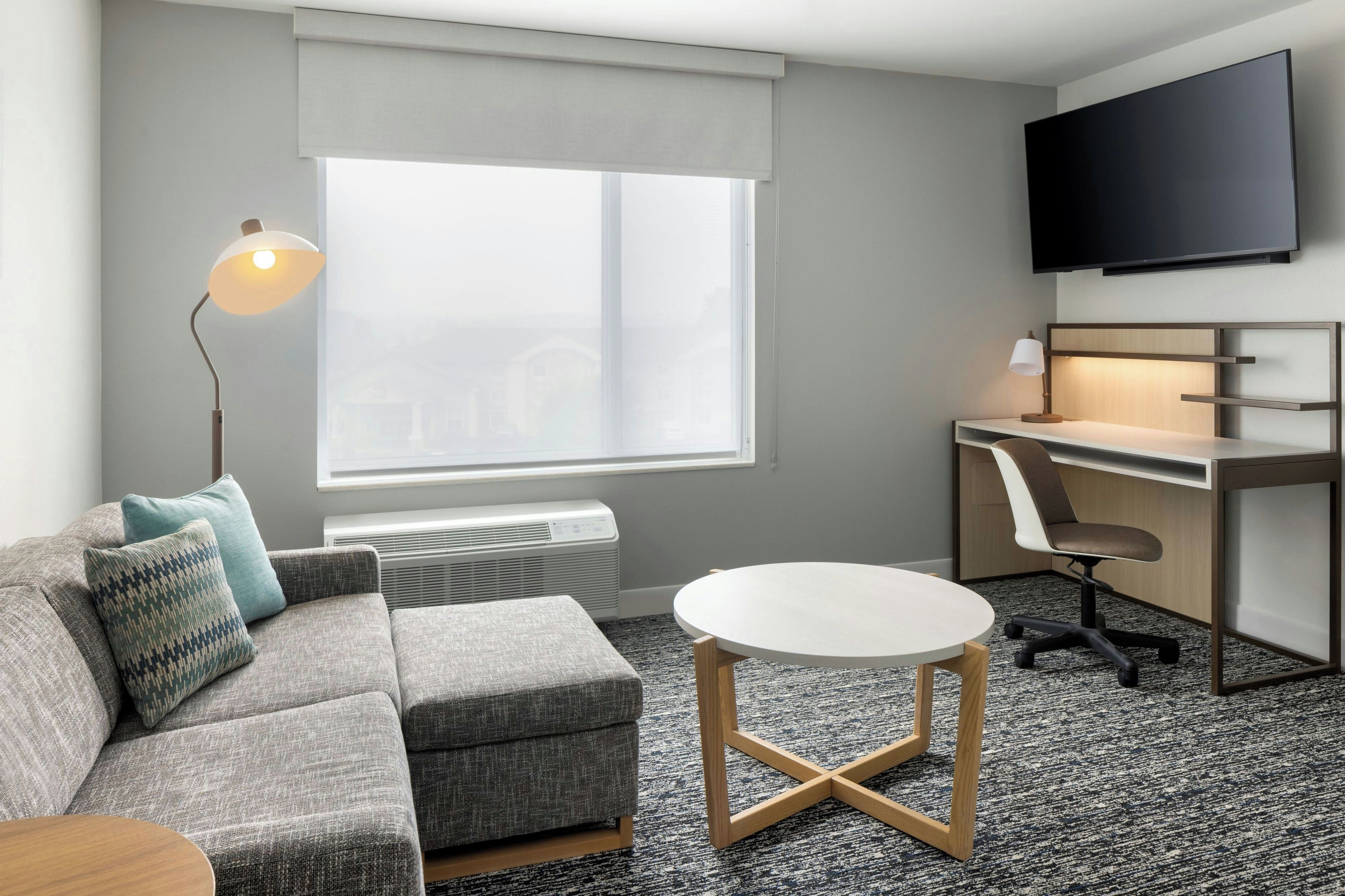 A modern hotel room with a grey sofa, a round table, a desk with a chair, a flat-screen TV, and a large window.