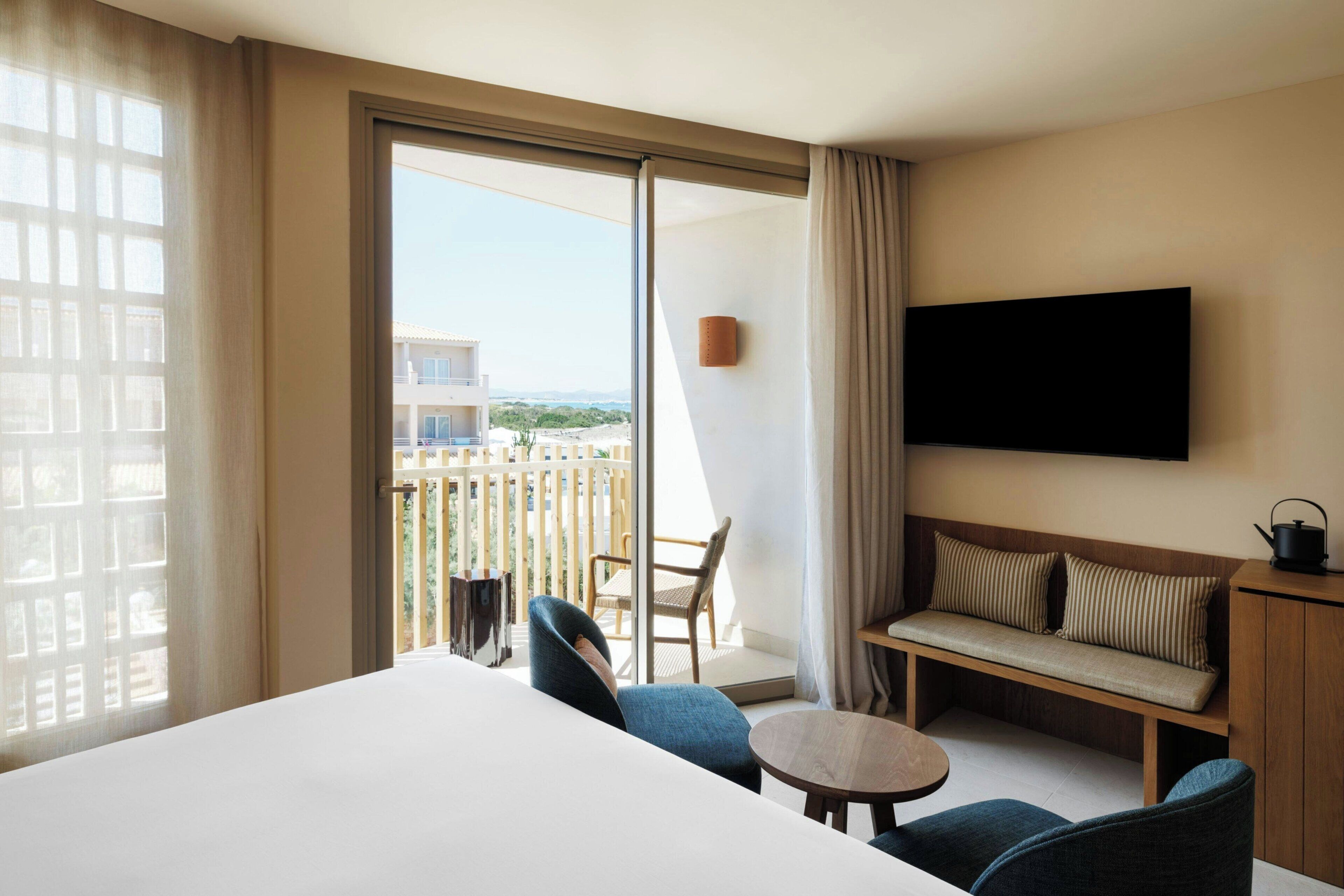 Hotelkamer met een groot bed, een bank, een tafeltje, een televisie en een balkon met uitzicht op gebouwen en groen.