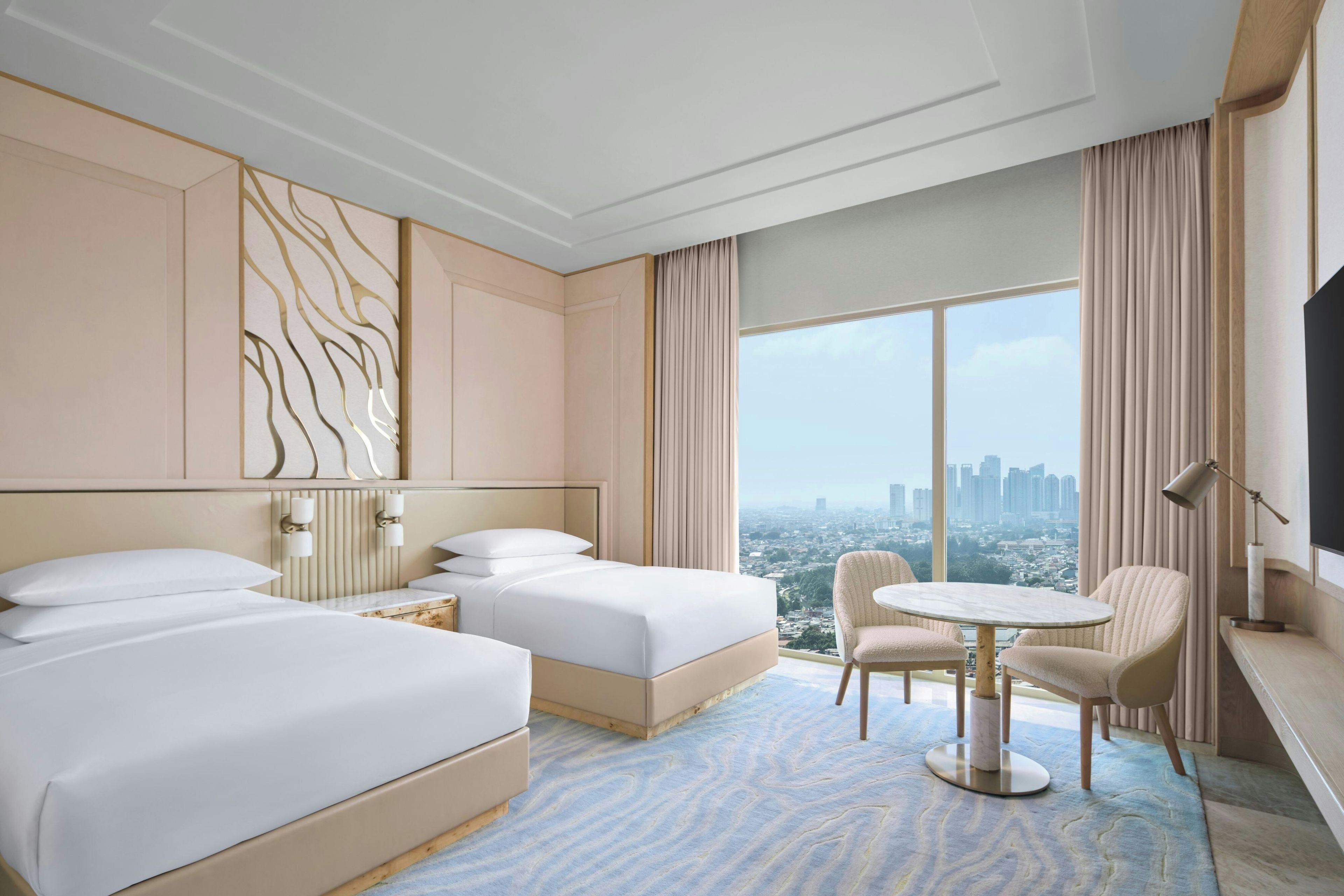 A hotel room with two beds, a sitting area with a table and chairs, a large window with a city view, and a mounted television.