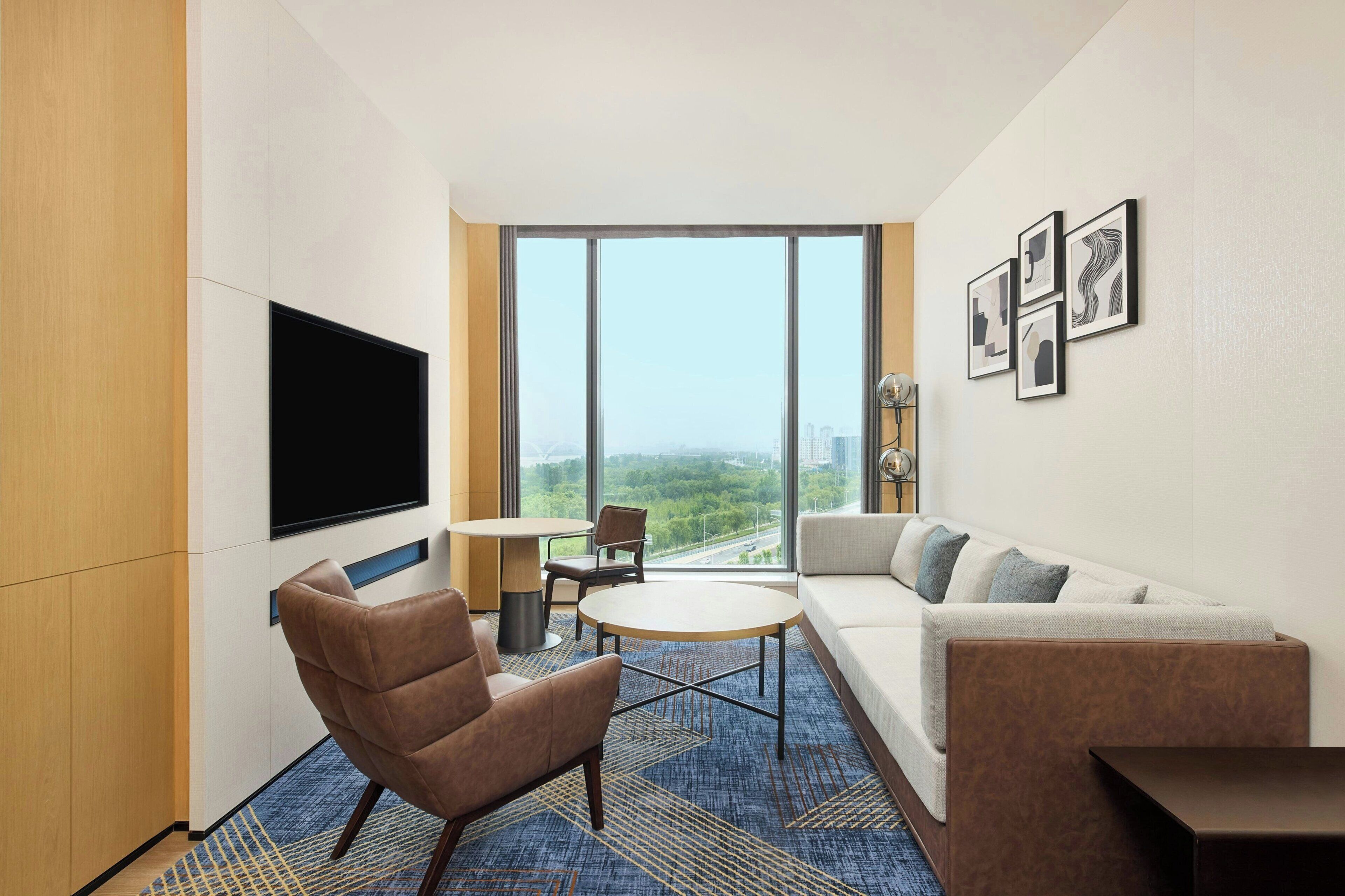 A modern hotel room with a large window, a flat-screen TV, a sofa, a chair, and a small table.