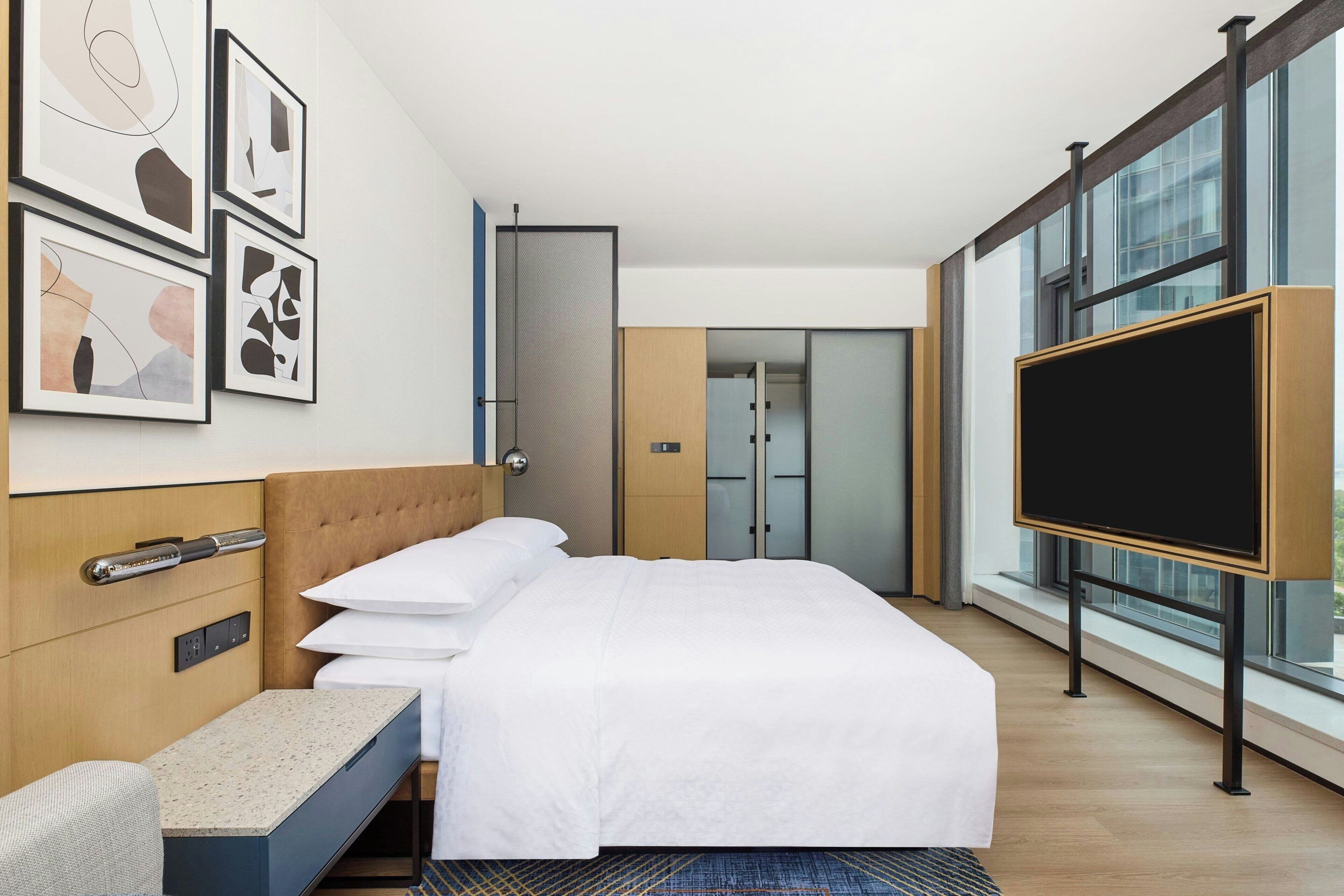 A modern hotel room with a bed, a desk, a flat-screen TV, and large windows.