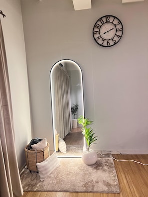 Ein Zimmer mit einem großen Spiegel, einer Uhr, einer Pflanze und einem kleinen Tisch mit Kissen.