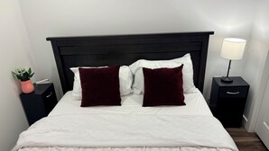 A bed with white bedding, two dark pillows, a black headboard, a bedside table with a lamp, and a small potted plant. - Victorious Place(STRA-2025-2882840) (Winnipeg)