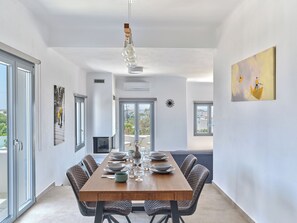 A modern dining area with a wooden table set for six, a chandelier, and artwork on the walls. - Mouragio Stay (Aliki)