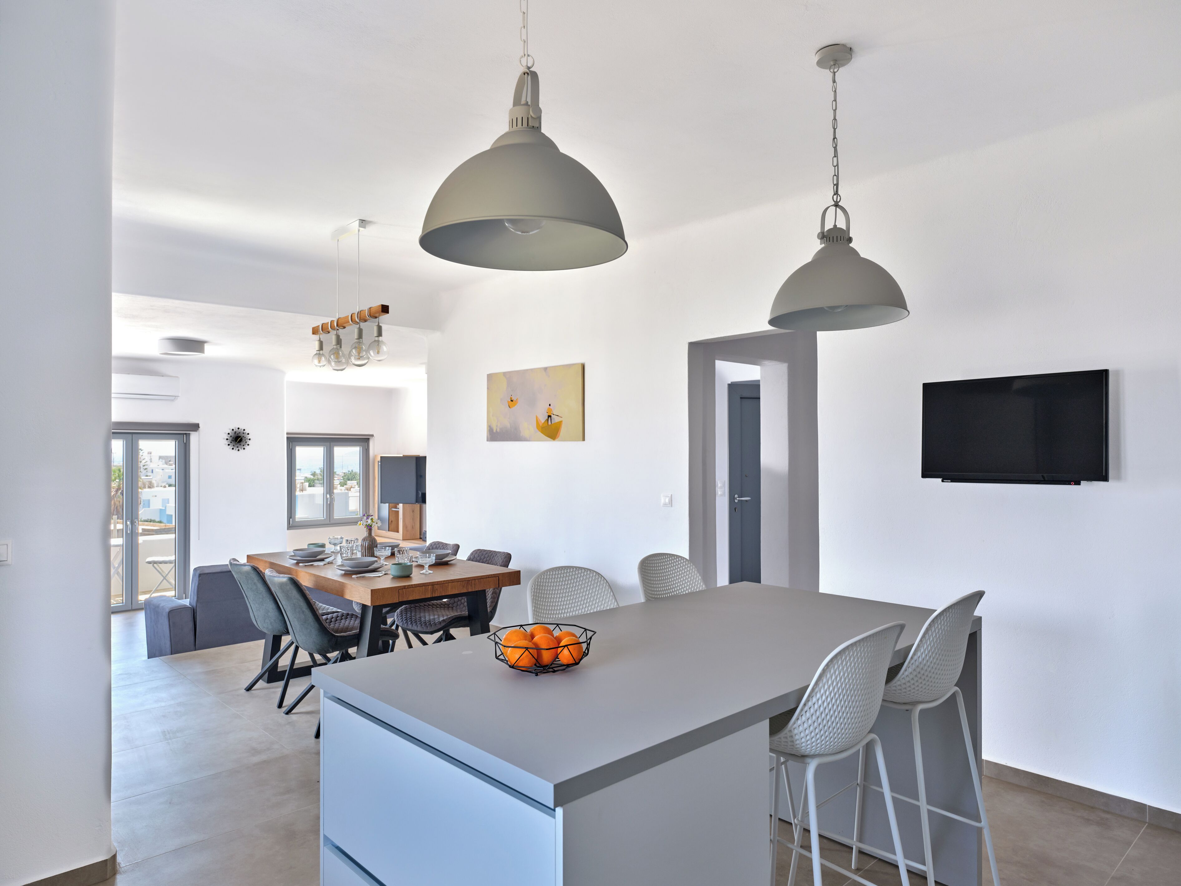 Een moderne keuken met een centraal eiland, hangende lampen, een eethoek met stoelen, een aan de muur bevestigde televisie en een deur naar een andere kamer.