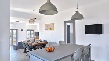 Een moderne keuken met een centraal eiland, hangende lampen, een eethoek met stoelen, een aan de muur bevestigde televisie en een deur naar een andere kamer.