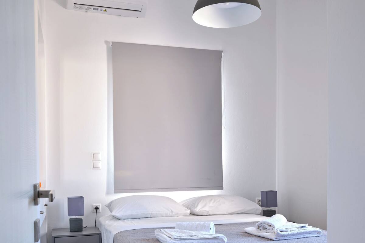 Un dormitori minimalista amb un llit, taules de nit, una làmpada penjant i un aparell d'aire condicionat.