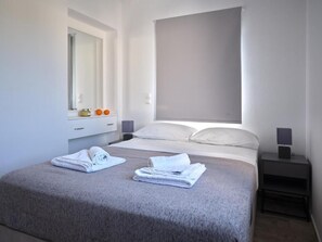 A modern bedroom with a large bed, a bedside table with a lamp, a window with a blind, and a mirror above the bed. - Mouragio Stay (Aliki)