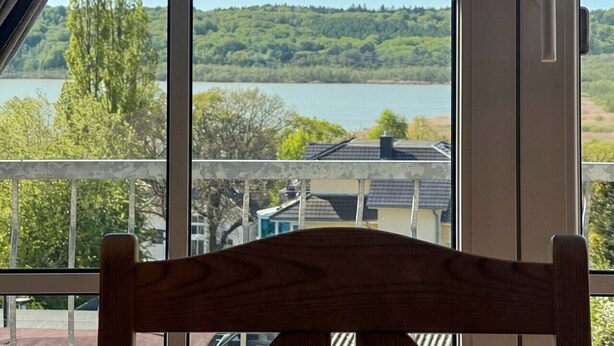 A room with a large window offering a view of a lake and hills. - Haus Seeblick - Panorama Suite No. 5 (Binz)