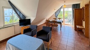 A room with a sloped ceiling, a dining area, a TV, and a view through the window. - Haus Seeblick - Panorama Suite No. 5 (Binz)