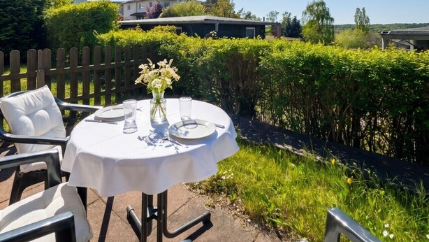 An outdoor dining area with a table set for two, surrounded by chairs, and a view of a house and greenery in the background. - Haus Seeblick - Comfort Apartment No. 2 with lake view (Binz)