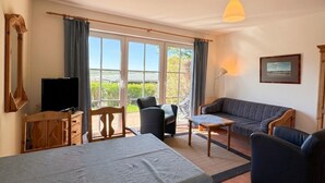 A room with a dining table, chairs, a sofa, a television, and a window with curtains. - Haus Seeblick - Comfort Apartment No. 2 with lake view (Binz)