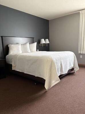 A hotel room with a bed, a nightstand, a lamp, and a window with curtains. - Prairie Inn of Jamestown (Jamestown)
