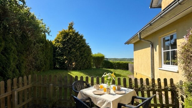 A patio with a table set for two, surrounded by a wooden fence, with a house in the background. - House Seeblick - Garden Suite No. 4 (Binz)