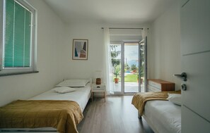 Ein Schlafzimmer mit zwei Betten, einem Nachttisch, einer Kommode, einem Fenster mit Jalousien und einer Tür, die auf einen Balkon führt.