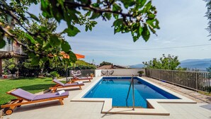 Ein rechteckiger Pool mit Blick auf Berge im Hintergrund, umgeben von einer eingezäunten Terrassenfläche mit Liegestühlen und einem kleinen Tisch.