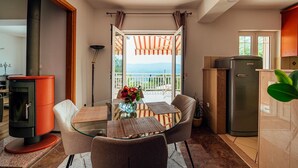 Ein Essbereich mit einem Glastisch, Stühlen, einer Vase mit Blumen, einer Küche mit Kühlschrank und Blick ins Freie.