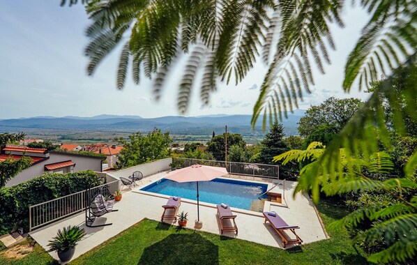 Ein Swimmingpool mit Liegestühlen, umgeben von viel Grün und Blick auf ferne Berge.