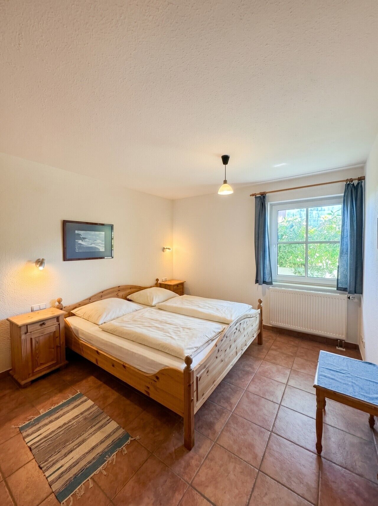 Ein Schlafzimmer mit Doppelbett, Nachttischen, einem Fenster mit Vorhängen, einem Heizkörper und einer Pendelleuchte.
