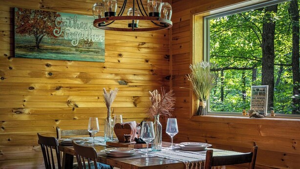 A wooden interior with a dining table set for six, a chandelier, and a window with a view of trees. - Peaceful 4-bedroom cabin with AC in charming Cosby (Cosby)