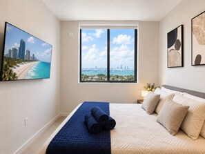 Ein Schlafzimmer mit Bett, einem Fenster mit Blick auf die Stadt, einem Strandbild und einer Lampe auf dem Nachttisch.