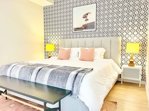 Ein Schlafzimmer mit einem Bett, Nachttischen, einer Bank, ornamentalem Wandpapier und einem gerahmten Bild an der Wand.