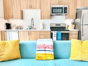 Eine Küchenzeile mit Mikrowelle, Backofen und Kühlschrank. Ein blaues Sofa mit gelben Kissen.