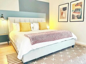 Ein ordentlich bezogenes Bett mit weißer Decke und einem gemusterten Überwurf, eine Nachttischlampe sowie zwei gerahmte Kunstwerke an der Wand.