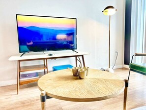 Ein modernes Wohnzimmer mit Fernseher auf einem Schrank, einem runden Holztisch mit Metallbechern, einer Stehlampe und einem grünen Sessel.