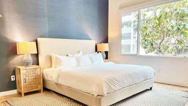 A bedroom with a bed, two bedside tables with lamps, a window with a view of trees, and a patterned rug. - King Bed Gaslamp, Beaches & Padres Nearby (San Diego)