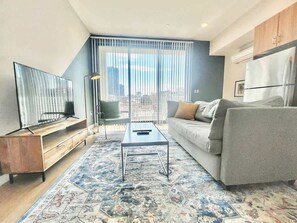 Ein modernes Wohnzimmer mit einer Couch, einem Couchtisch, einem Fernseher und einem großen Fenster mit Jalousien.