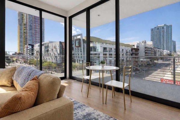 A modern living room with a large window, a sofa, a small table, and chairs. - Modern Downtown Retreat | Balcony + Rooftop Views (San Diego)