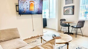 Ein Wohnzimmer mit einer Couch, einem Couchtisch, einem an der Wand befestigten Fernseher, einem Esstisch mit Stühlen und einem großen Fenster mit Blick auf ein Gebäude.