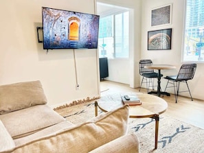 A living room with a sofa, a coffee table, a TV mounted on the wall, a dining table with chairs, and a large window with a view of a building. - Bars, Eats & Nightlife | Central San Diego King Apt (San Diego)