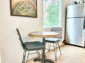 A dining area with a round table and two chairs, a refrigerator, and a window with a view of greenery. - Bars, Eats & Nightlife | Central San Diego King Apt (San Diego)