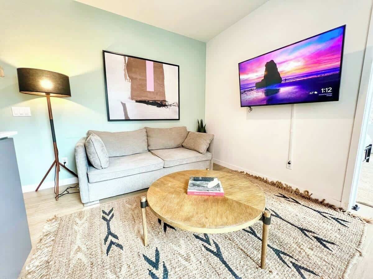 A living room with a sofa, a coffee table with books, a lamp, a TV on the wall, and a patterned rug.