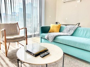 Ein modernes Wohnzimmer mit einem türkisfarbenen Sofa, einem Holzstuhl, einem runden Couchtisch mit Buch und Fernbedienung und Blick auf die Stadt durch große Fenster.