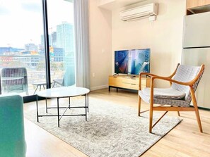 Ein modernes Wohnzimmer mit großem Fenster, einem Flachbildfernseher, einem Holzstuhl, einem kleinen Tisch und einer Küchenzeile.