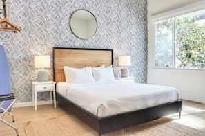 Una camera da letto con un letto, due comodini, uno specchio rotondo e una finestra vista verde.